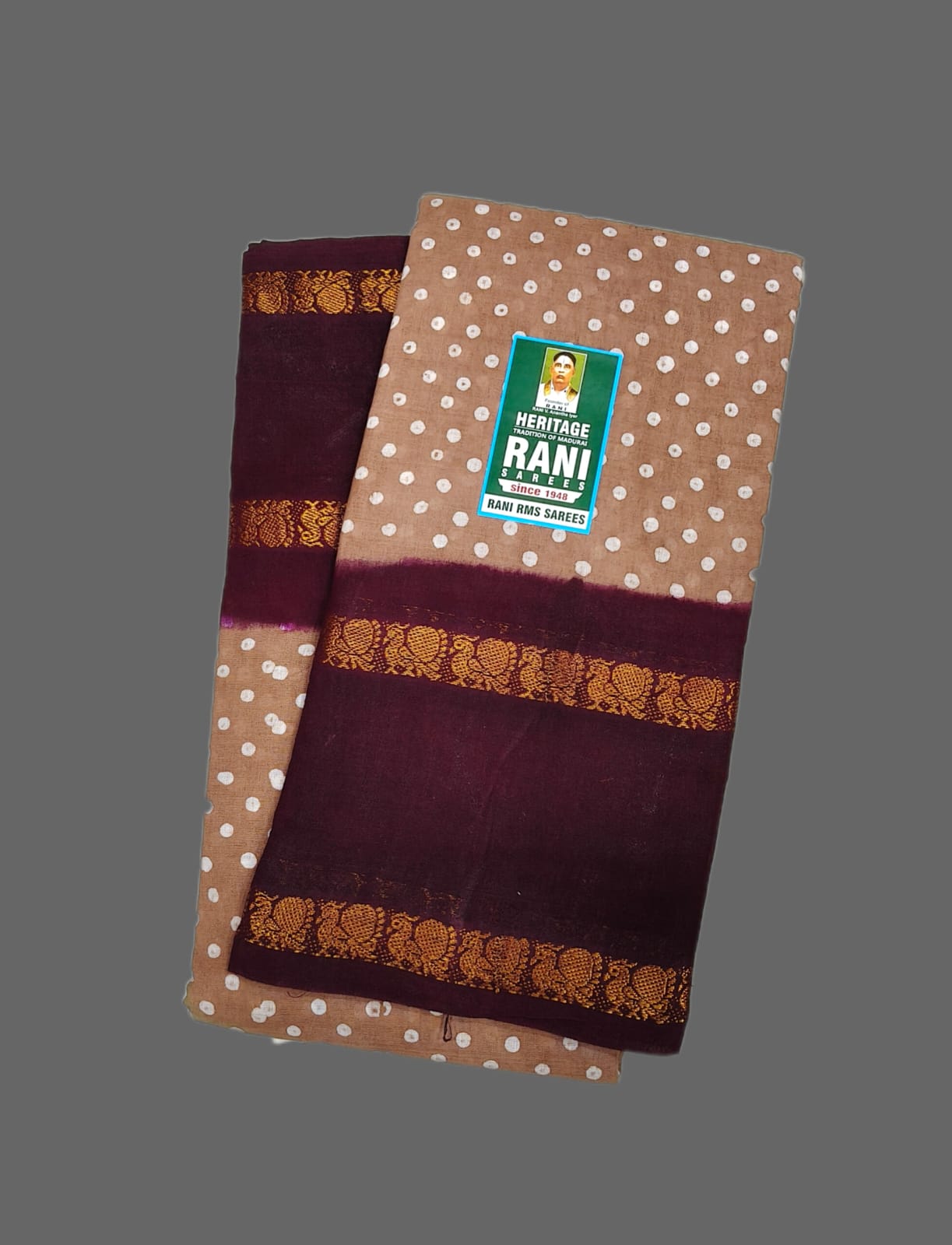 A folded Heritage Rani Handloom cotton fabric piece, likely a sari, in brown with white polka dots, deep maroon borders, and gold zari motifs, displayed on a solid gray background.